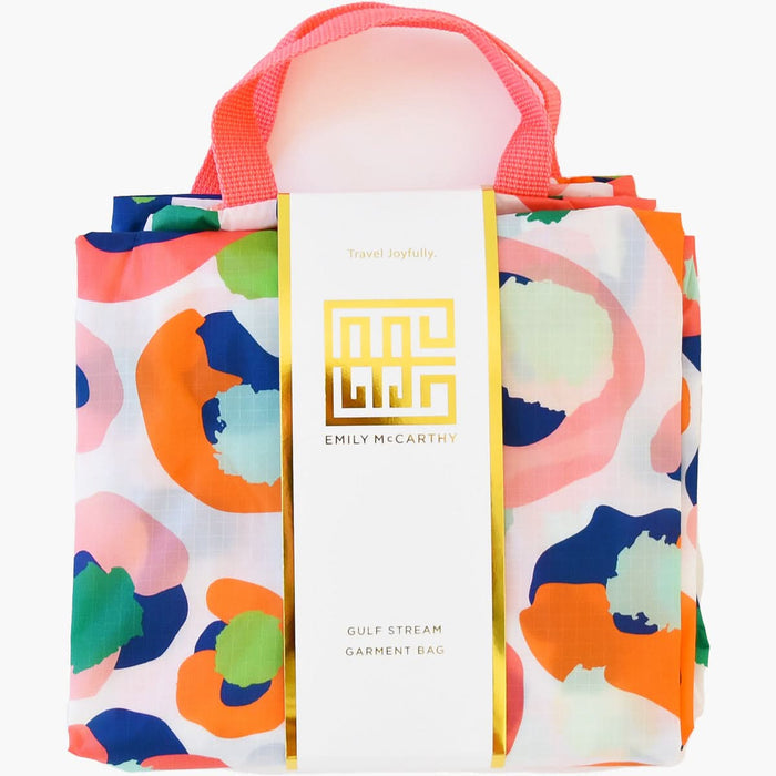 The Gulf Stream Garment Bag - Multi Spot Cheetah from the Emily McCarthy Collection showcases a vibrant abstract pattern in blue, green, pink, and orange. Featuring a pink handle and a gold label that reads "Emily McCarthy" and "Travel Joyfully," this bag combines style with practicality for your adventures.
