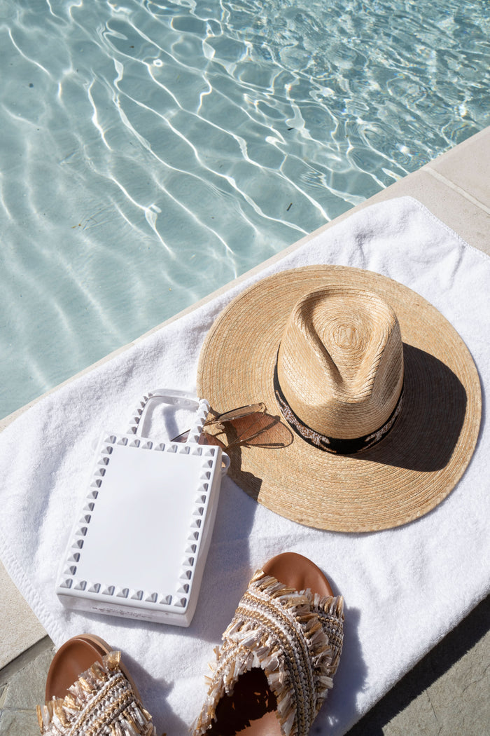 A Sunshine Tienda x Emily McCarthy Chasing Cheetah Hat, a white handbag, and tassel-adorned sandals rest on a white towel beside a clear swimming pool, completing the perfect swimwear ensemble for a sunny day.