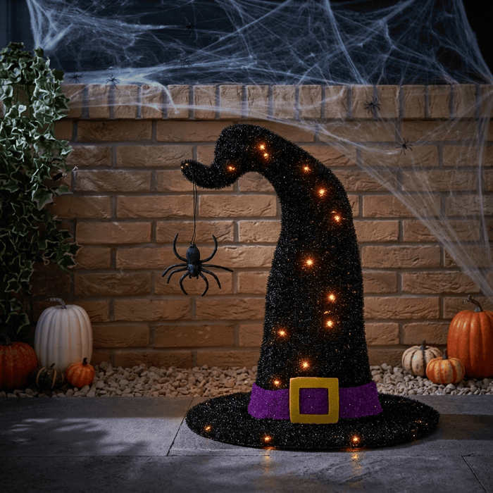 A glittery black witch hat Halloween decoration with a purple band, gold buckle, and warm glowing lights, displayed in a spooky outdoor patio setting with pumpkins, cobwebs, and a spider.