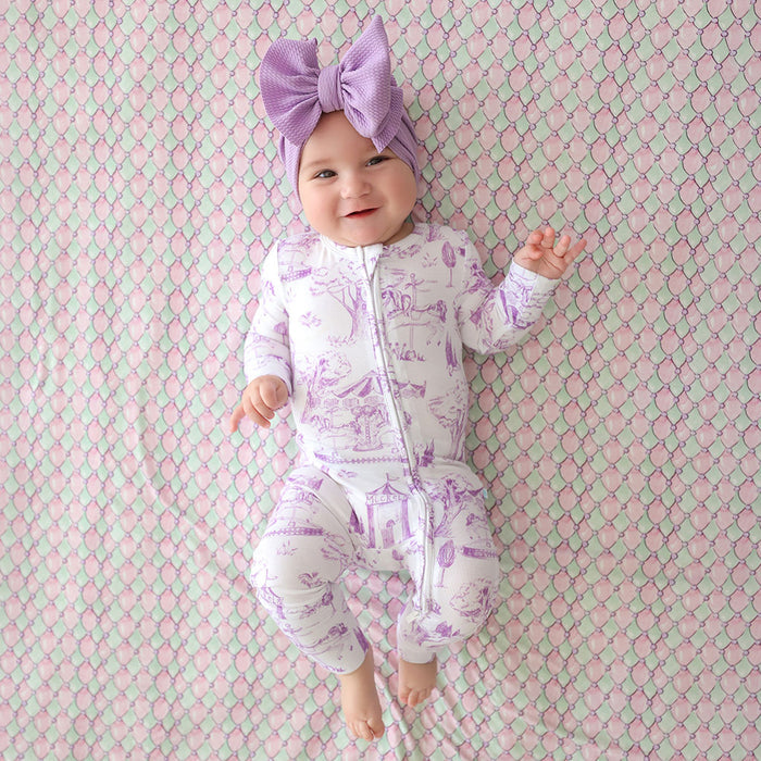 Baby in a purple and white outfit with a matching headband on a patterned pink and green background