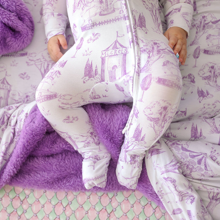 Child in a purple and white patterned onesie sitting on a purple blanket.