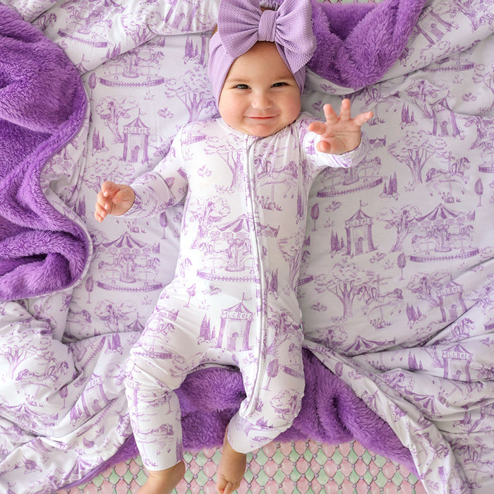 Baby in a white onesie with purple patterns, surrounded by purple blankets and headband on a patterned surface.