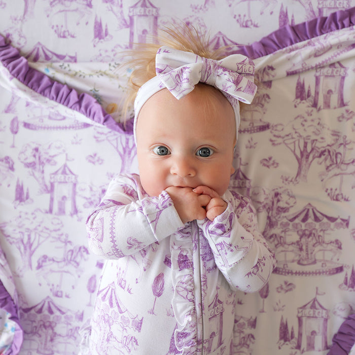 Baby in a purple and white patterned outfit with a matching headband, sitting on a purple and white patterned surface.