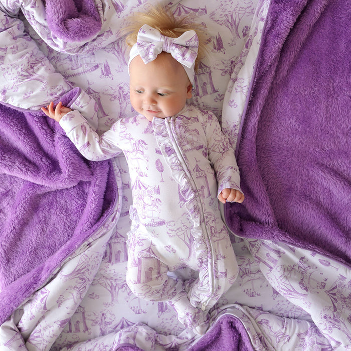 Baby lying on a purple plush blanket wearing a matching one piece.