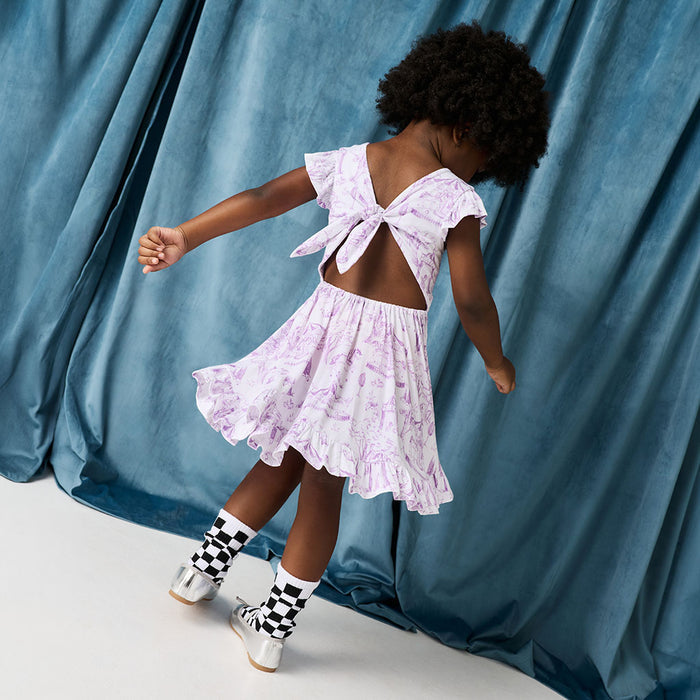 Child wearing a light purple dress with a back bow, standing against a blue curtain.