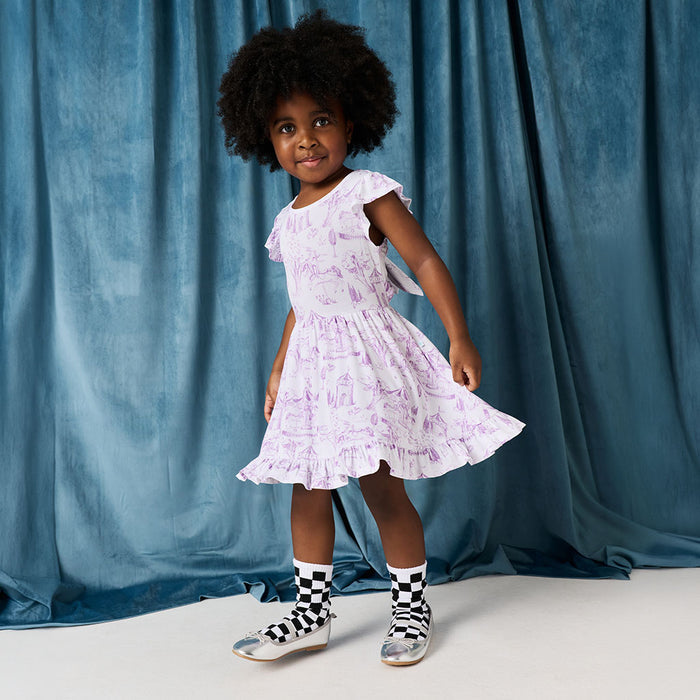 Young girl in a white dress with purple floral pattern standing against a blue curtain.