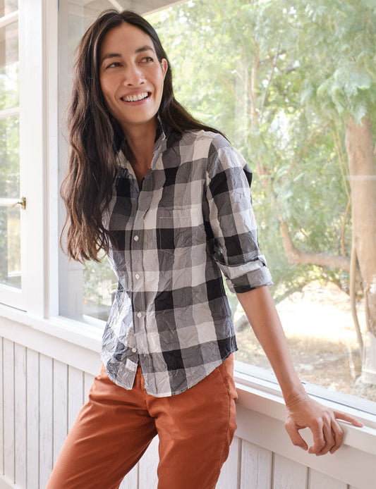 A woman smiling while wearing a black and white checkered button-down shirt and rust-orange pants, leaning on a white porch railing with trees visible in the background.