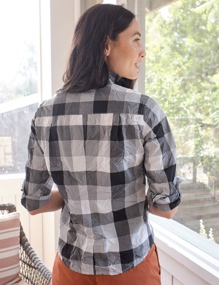 A woman wearing a black, white, and gray buffalo plaid button-down shirt, shown from the back as she looks out a window.