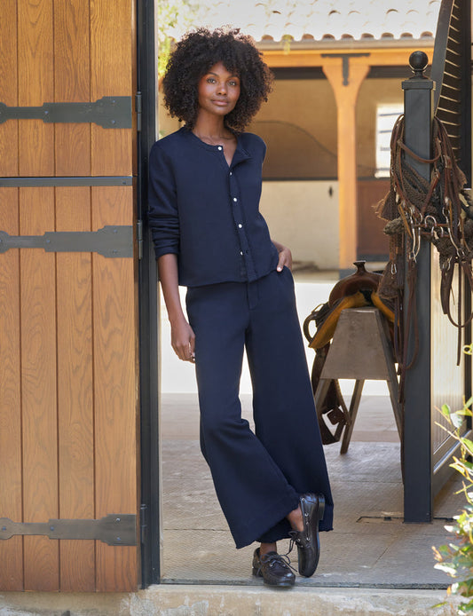 A woman with curly hair stands in a doorway wearing a navy blue long-sleeve cardigan and matching wide-leg pants, paired with black shoes. The setting appears to be a rustic indoor-outdoor space with wood paneling, a saddle, and warm architectural details in the background.