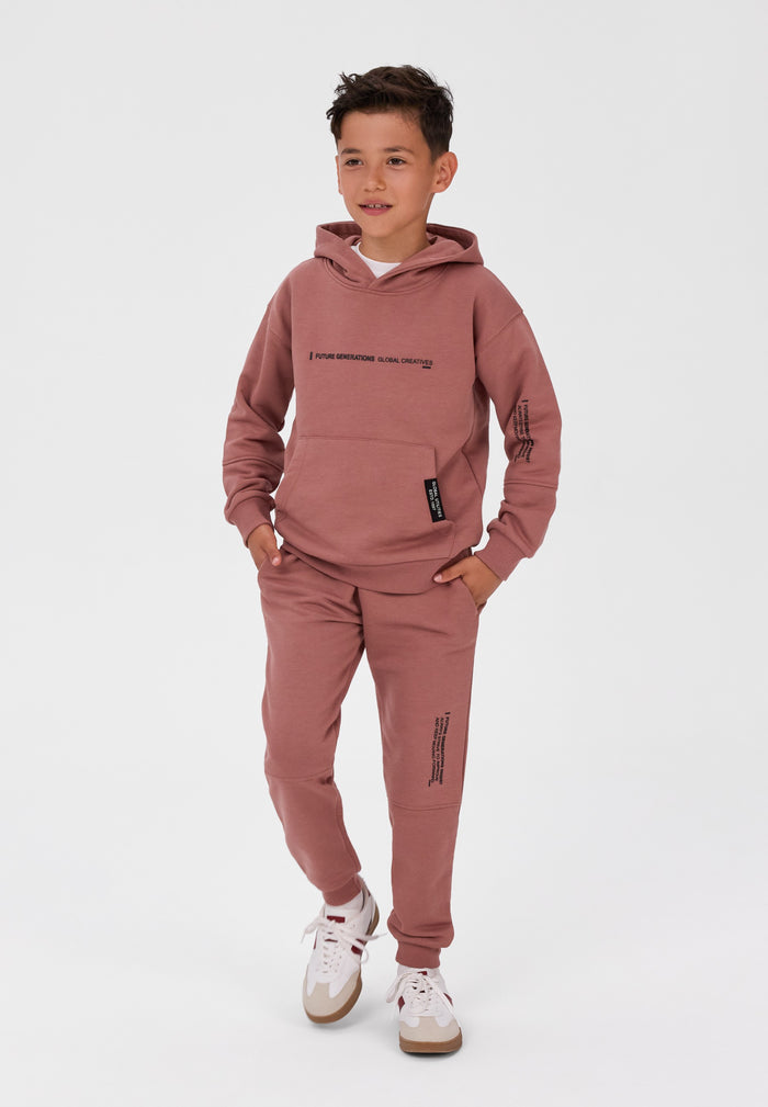 A young boy modeling a dusty rose pink two-piece tracksuit, featuring a pullover hoodie with black graphic text on the chest and sleeve, matching jogger pants with vertical text graphics, and white sneakers, set against a plain light gray studio background.