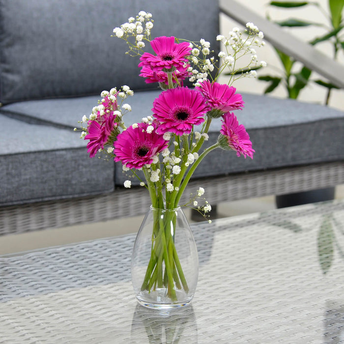 Clear glass flower vase filled with pink gerbera daisies and gypsophila on a glass table with grey sofa