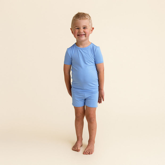 Boy standing wearing the Posh Peanut Chatham Blue Classic Pajama Short Set, a solid blue short-sleeve top and shorts.