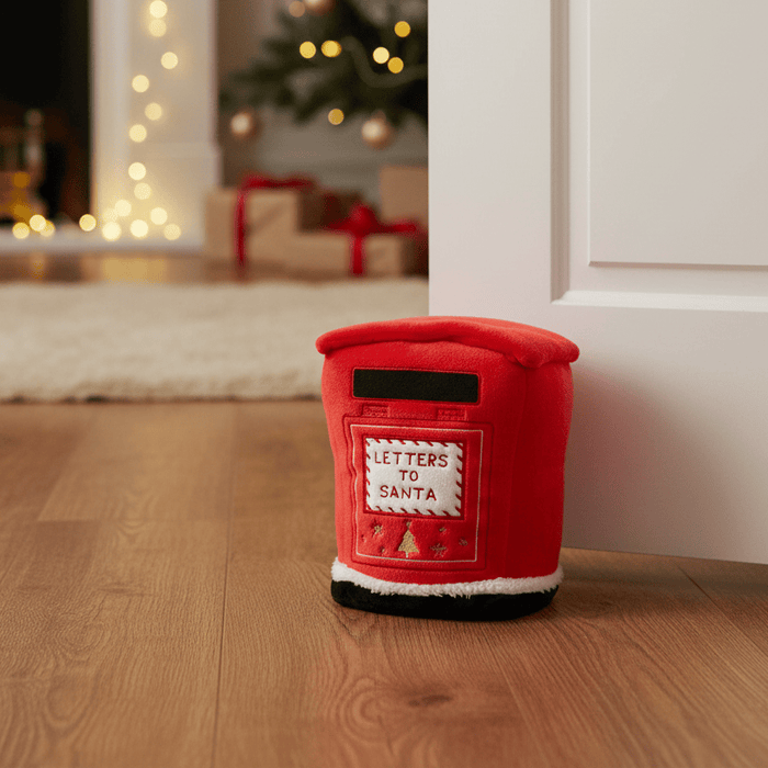 A red plush postman-shaped door stopper with a front patch reading 'LETTERS TO SANTA' sits on a wooden floor in a doorway, with a blurred Christmas tree and fireplace in the background.