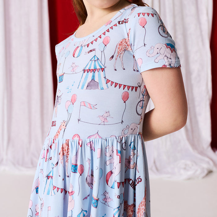 Child wearing a light blue dress with circus-themed print against a white curtain background