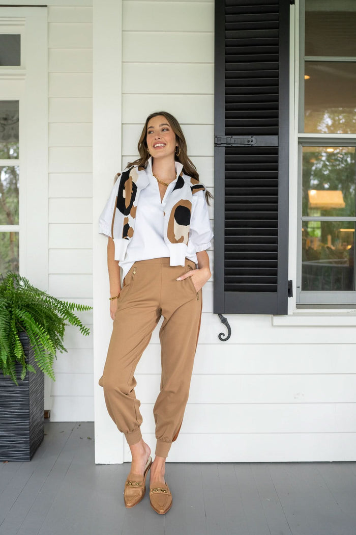 A woman in Downtown Jogger - Luggage Ponte pants and a white blouse with a patterned scarf stands on a porch. She is smiling and leaning against a white wall with black window shutters, while a potted fern sits nearby.