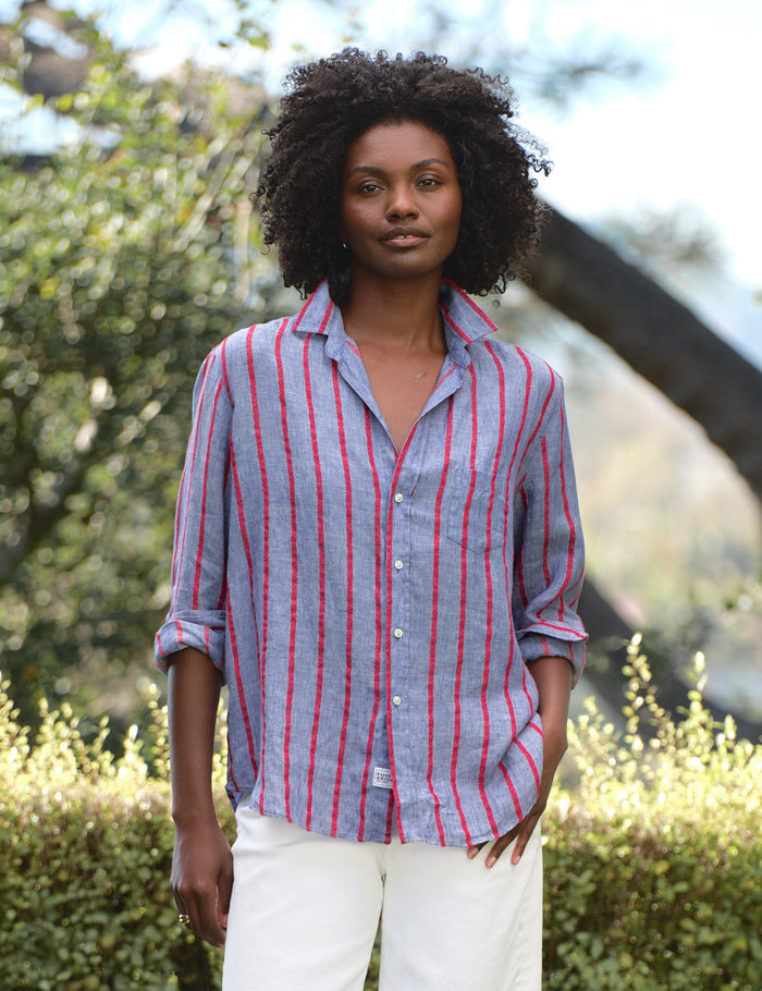 Woman wearing a striped shirt and white pants standing outdoors with greenery in the background