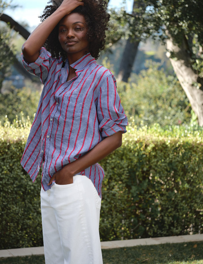 Woman wearing a red and blue striped shirt and white pants standing outdoors with greenery in the background