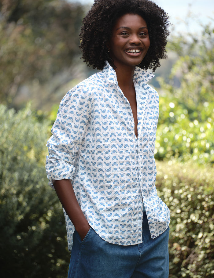 Woman wearing a white patterned shirt with rolled-up sleeves and blue jeans standing outdoors.