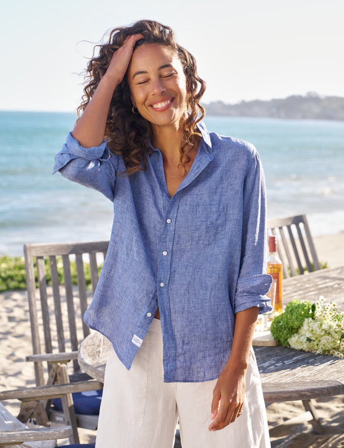 Woman wearing a blue shirt and white pants standing on a beach with ocean view.