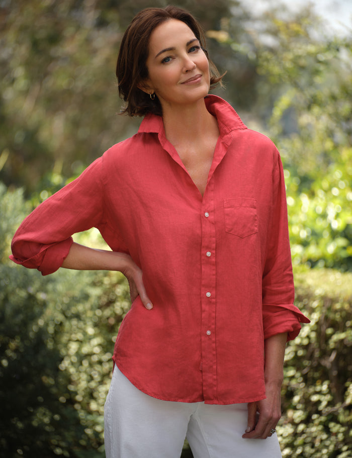 Woman wearing a red shirt and white pants standing outdoors with greenery in the background