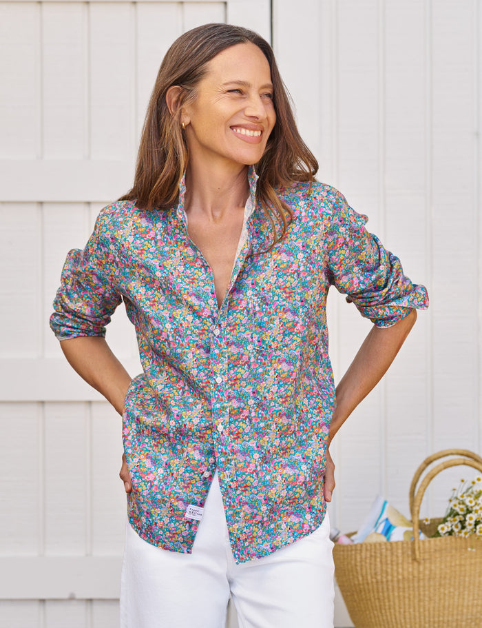 Woman wearing a colorful floral shirt with white pants standing against a light wooden wall.