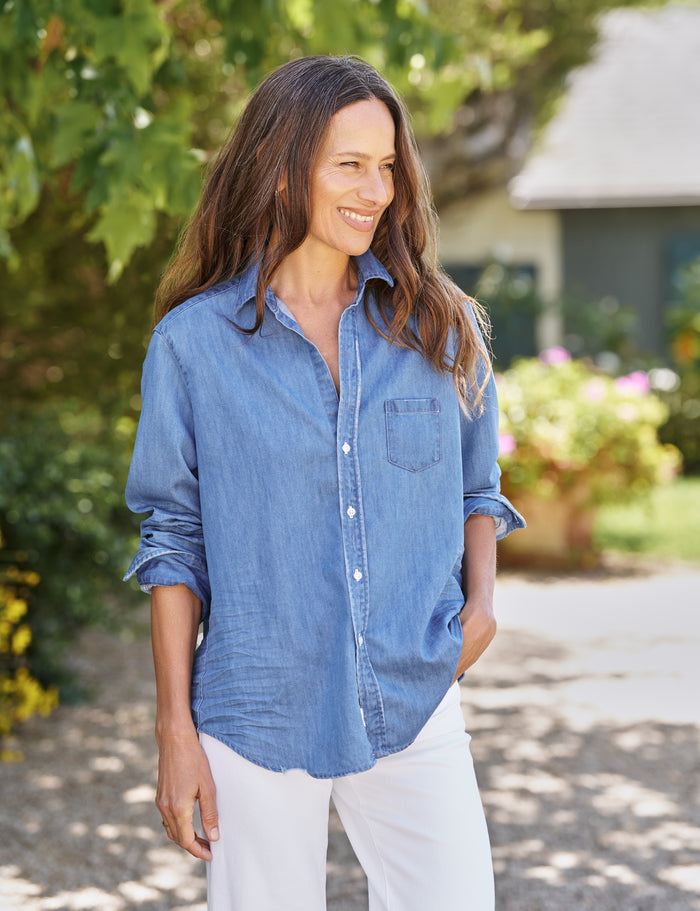 Woman wearing a blue denim shirt and white pants standing outdoors with greenery in the background