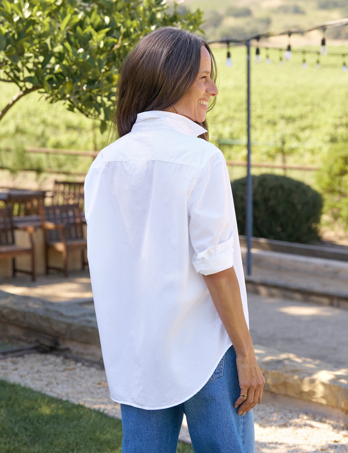 Woman wearing a white shirt and blue jeans standing outdoors with greenery in the background