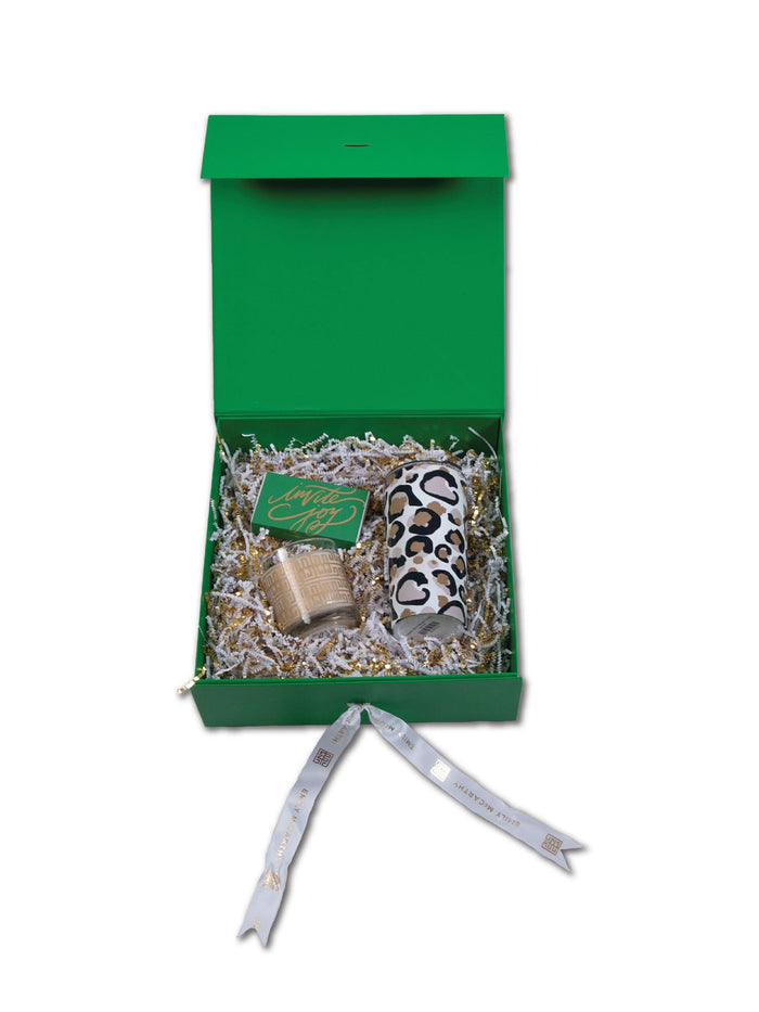 The Emily McCarthy Gift Box features a green box with shredded paper, a leopard-print insulated tumbler, a candle, a green card, and is elegantly finished with a white ribbon with gold accents draped in front.