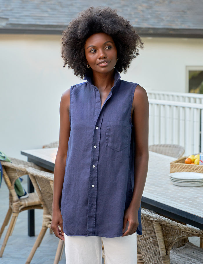 Woman wearing a sleeveless blue shirt standing outdoors near a table with chairs.