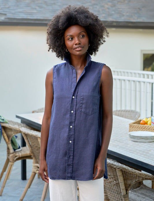 Woman wearing a sleeveless blue shirt standing outdoors near a table with chairs.