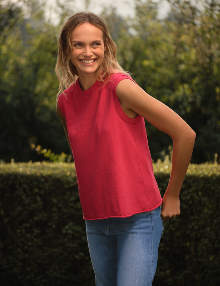 Woman wearing a red sleeveless top and blue jeans standing outdoors with greenery in the background