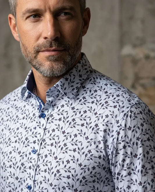 Man wearing a white shirt with a subtle blue floral pattern against a textured wall.