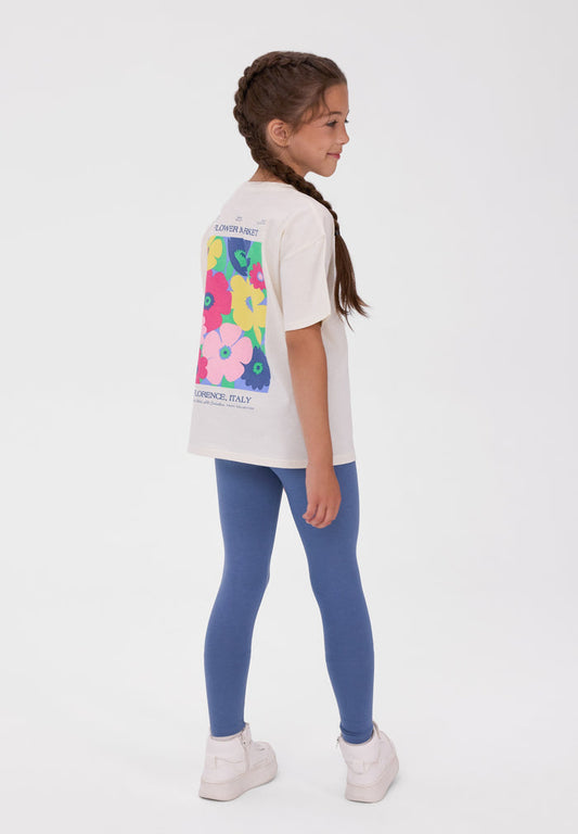 A young girl with a braided ponytail is shown from the back wearing a white t-shirt with a colorful floral graphic and blue leggings, standing against a plain white studio background.
