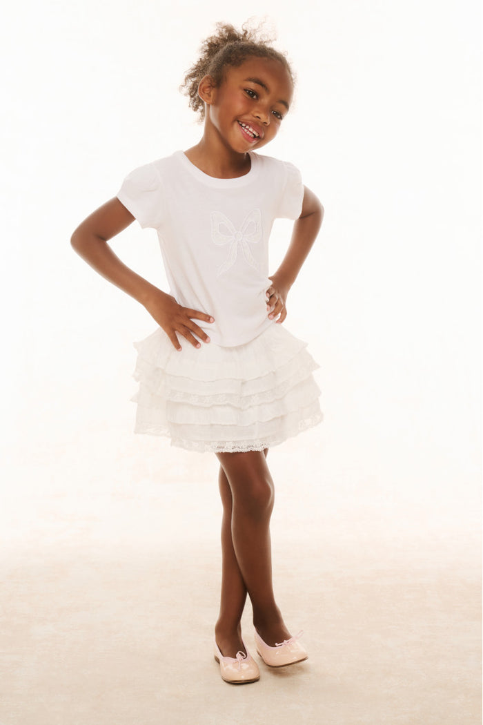 A young girl standing against a white background, wearing a white knee-length skirt with ruffle details and a wide smocked waistband, paired with a white t-shirt and white shoes.