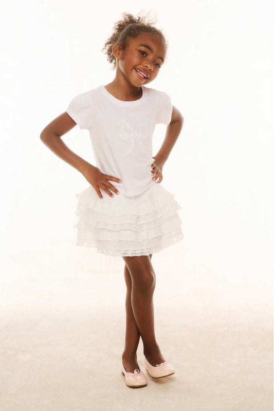 A young girl standing against a white background, wearing a white knee-length skirt with ruffle details and a wide smocked waistband, paired with a white t-shirt and white shoes.