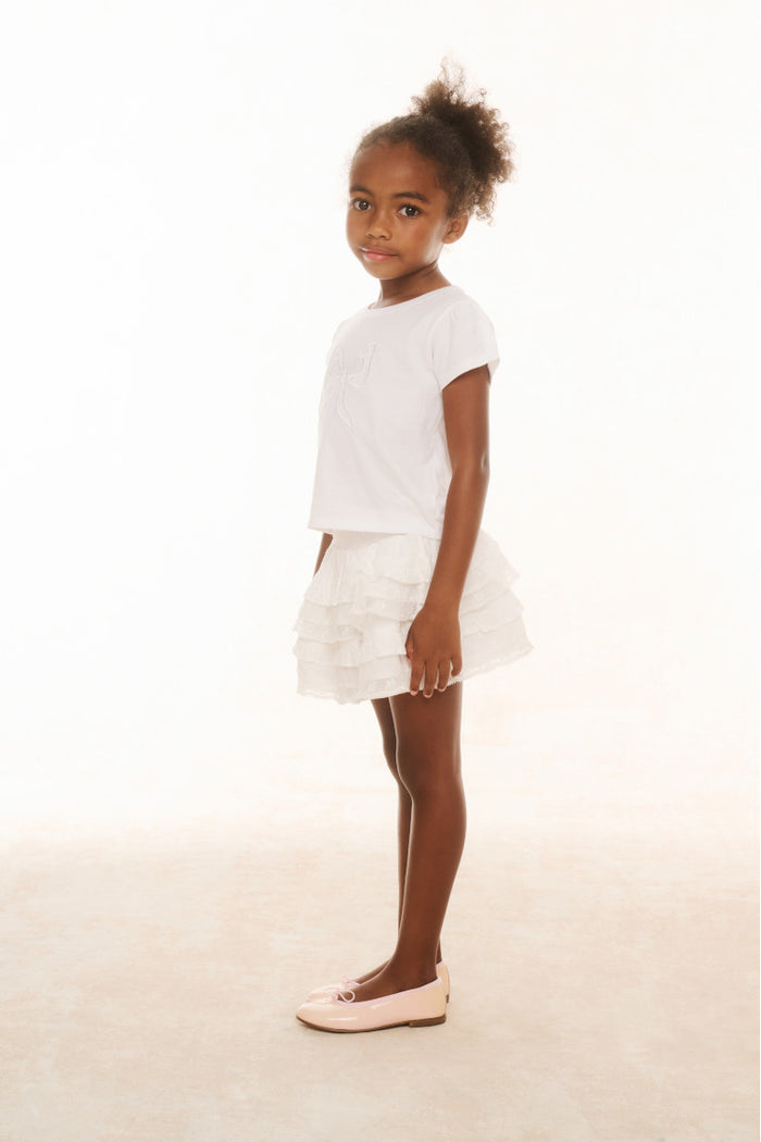 A young girl standing against a white background, wearing a white knee-length skirt with ruffle details and a wide smocked waistband, paired with a white t-shirt and white shoes.
