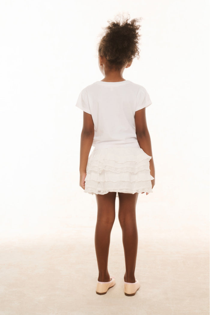 A young girl standing against a white background, wearing a white knee-length skirt with ruffle details and a wide smocked waistband, paired with a white t-shirt and white shoes.