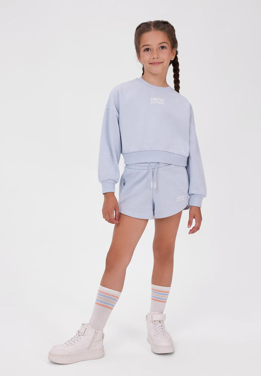 A young girl with braided hair stands against a light gray studio background wearing a light blue cropped sweatshirt and matching shorts, with white socks and white sneakers.