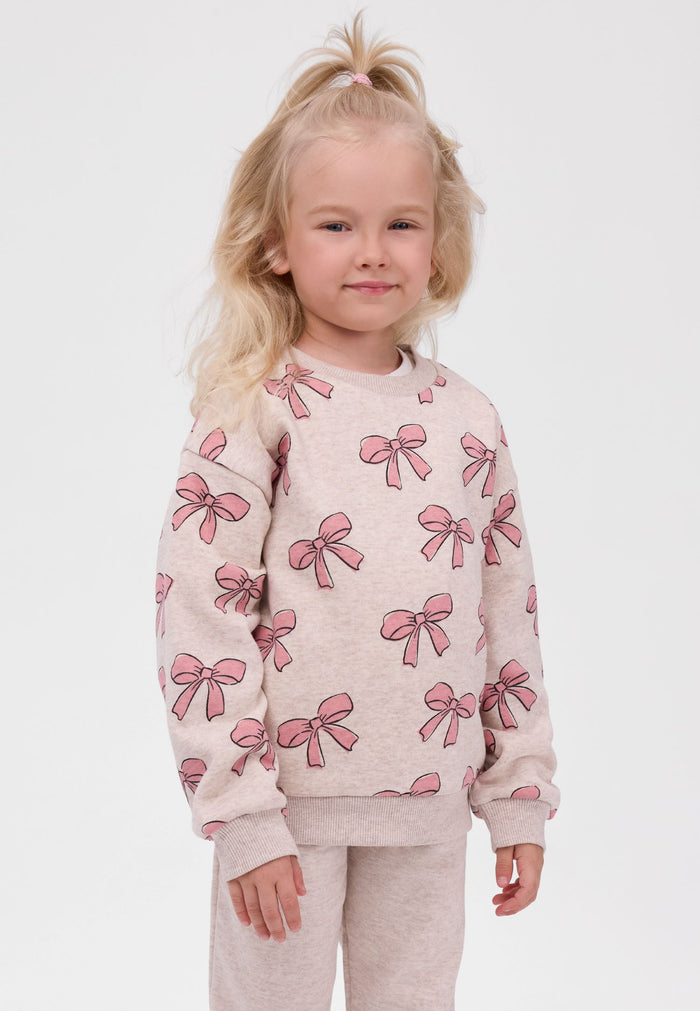 A young girl with blonde hair in a high ponytail wearing a light beige crewneck sweatshirt with an all-over pink bow pattern, paired with matching beige pants, against a plain light gray background.