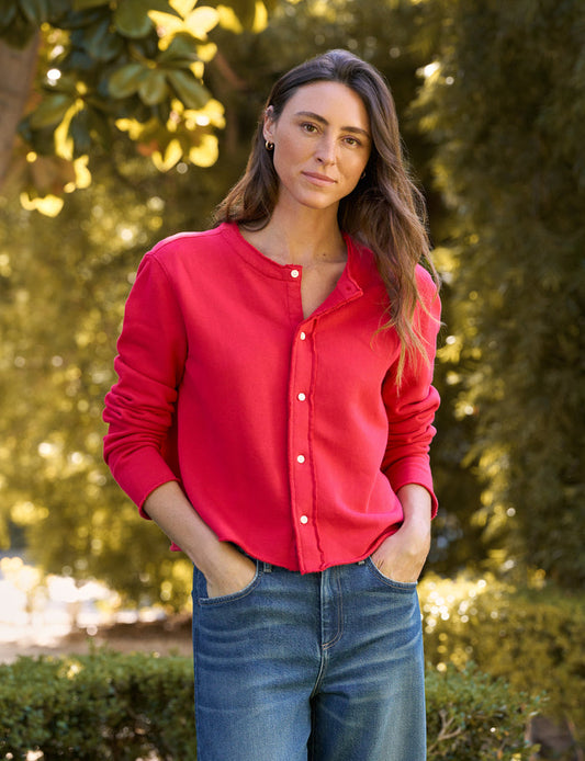 A woman wearing a bright red long-sleeve button-up shirt and blue jeans, standing outdoors in front of a blurred green garden background.