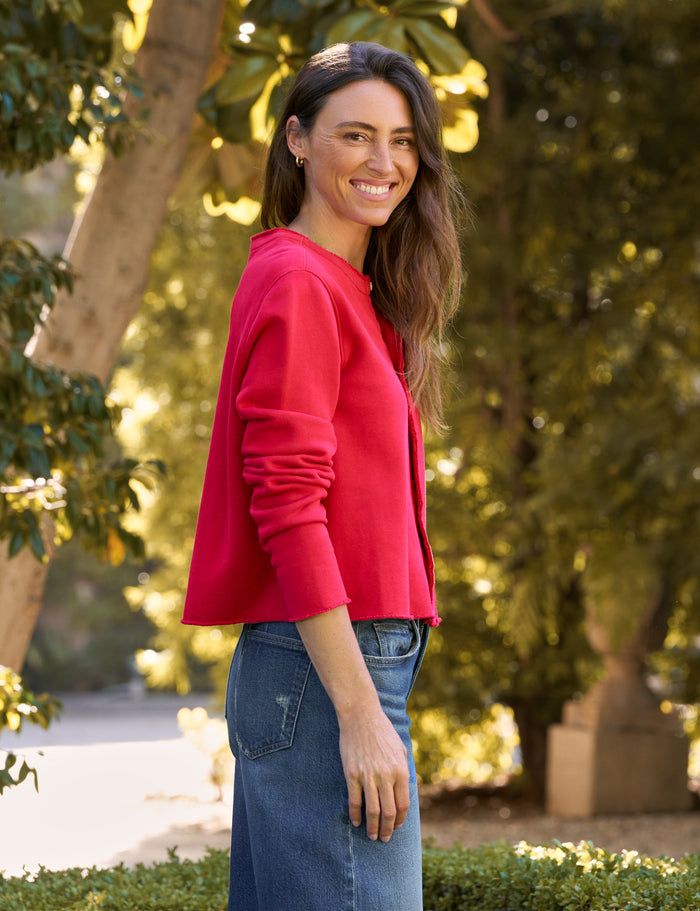 A woman wearing a bright red cropped long-sleeve top and blue jeans, standing outdoors in front of a softly blurred background of trees and foliage.