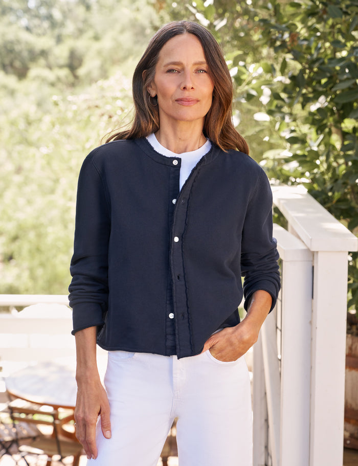 Woman wearing a navy cardigan over a white shirt and pants, standing outdoors with greenery in the background.