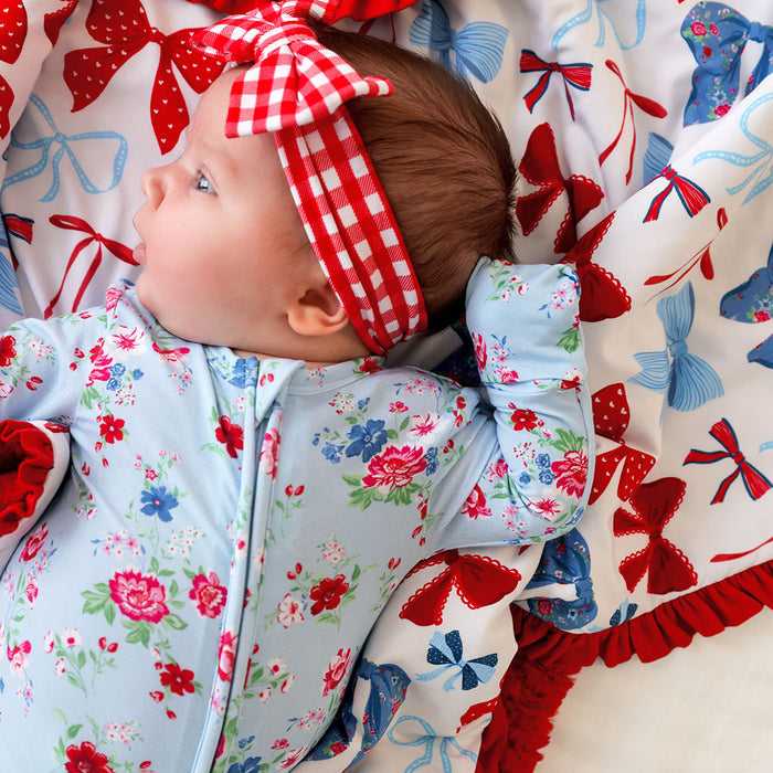 Baby wearing a floral outfit and red checkered headband on a blanket with red, white, and blue bows.