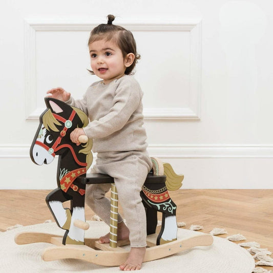 Child sitting on a wooden rocking horse in a room with white walls and wooden floor.