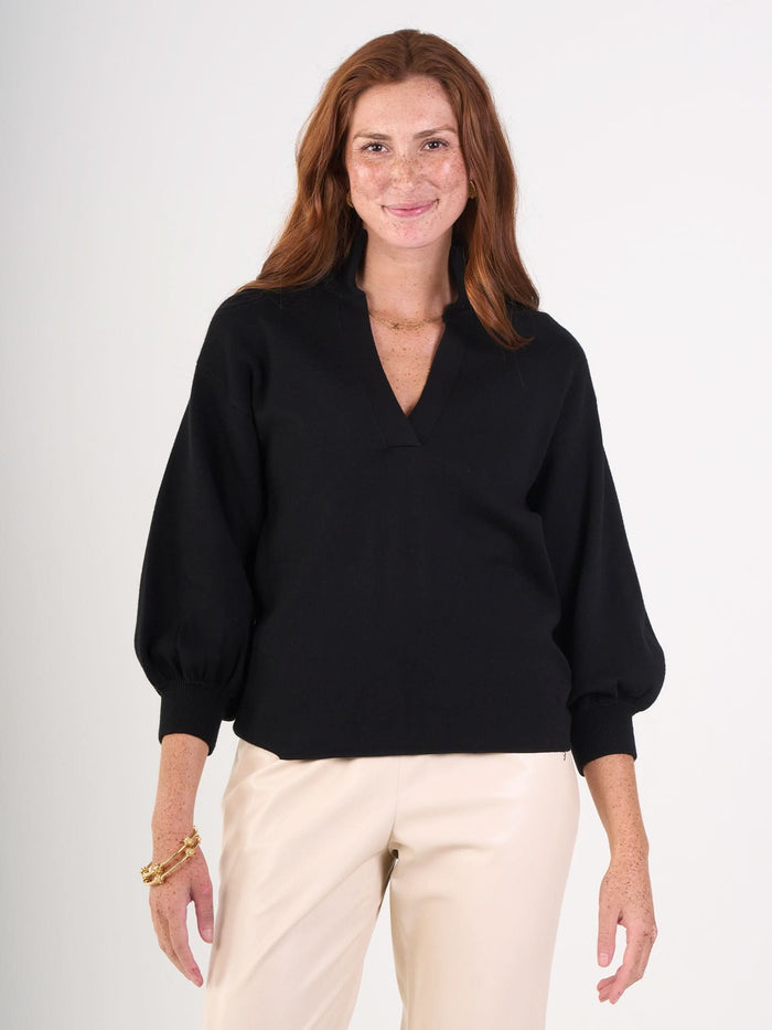 A smiling woman with long red hair wears the Lolli Sweater, a black viscose knit V-neck blouse with balloon sleeves, and cream-colored pants, standing against a plain white background.