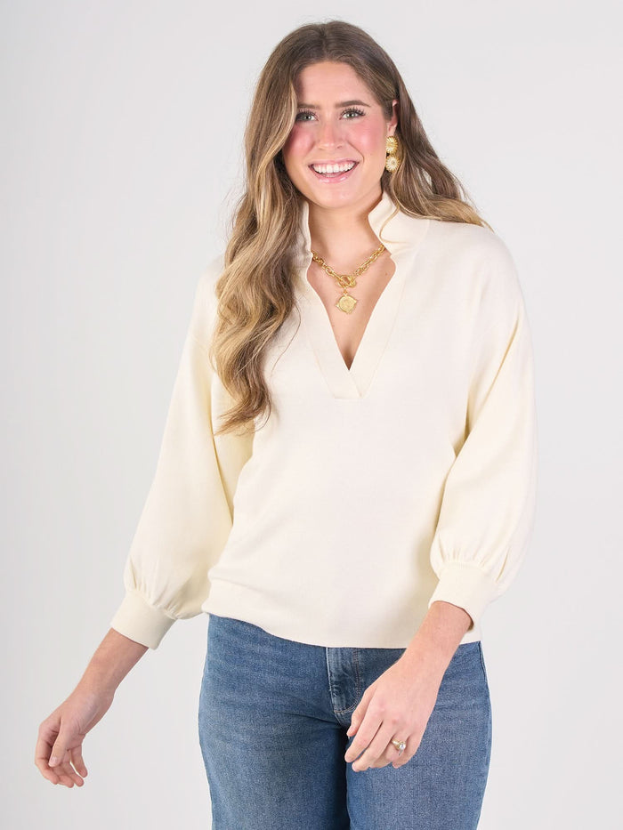 A woman with long, wavy hair wears the Lolli Sweater—a cream-colored blouse with blouson sleeves—paired with blue jeans and gold jewelry, smiling against a plain light background.
