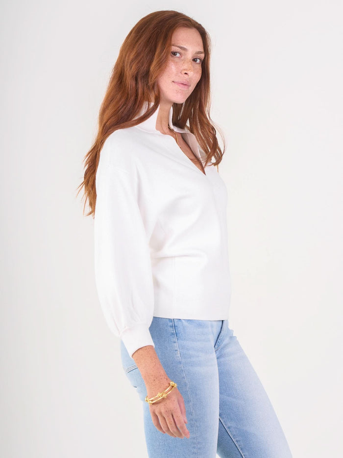A woman with long red hair models the white Lolli Sweater and light blue jeans, posing against a plain white background. She smiles slightly, her hand by her side showing a gold bracelet.