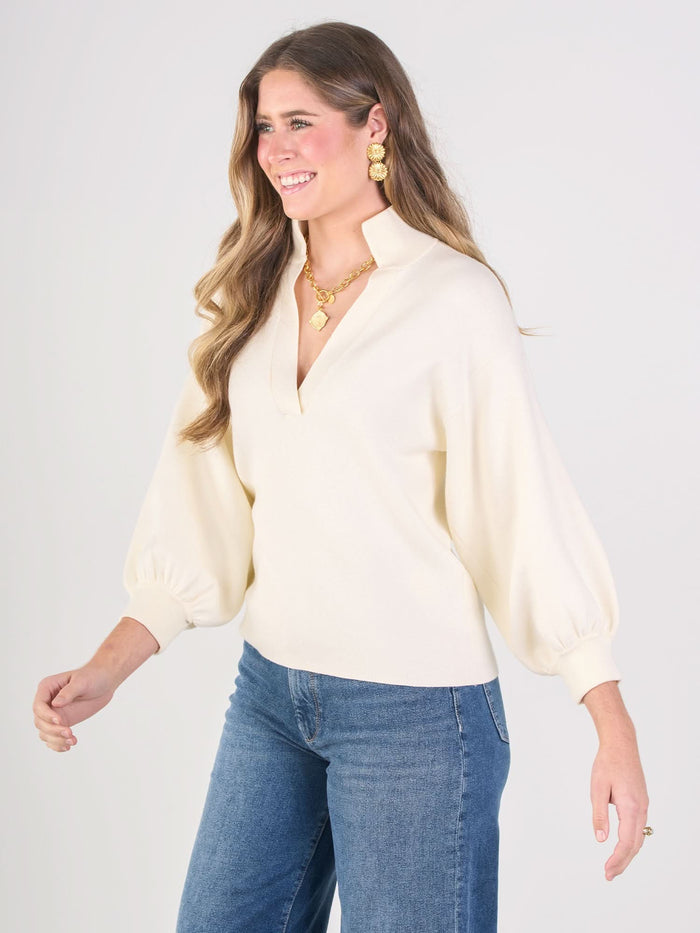 A woman with long wavy hair smiles in a cream Lolli Sweater with blouson sleeves, paired with blue jeans, gold earrings, and a gold necklace, standing against a plain light background.