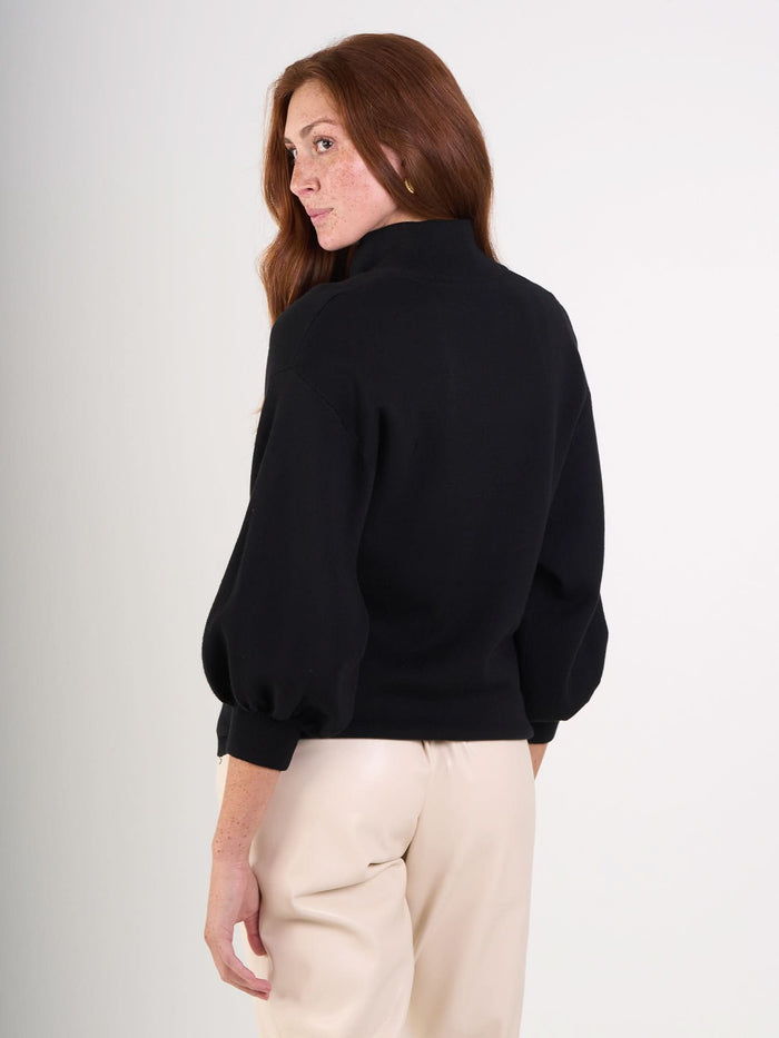 A person with long, auburn hair wears the black Lolli Sweater with puffed sleeves, made from viscose knit, paired with light beige pants, standing sideways and looking over their shoulder against a plain background.