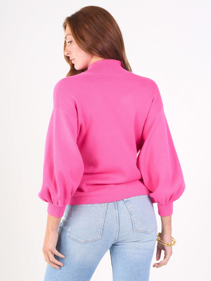 A woman with long red hair wears the Lolli Sweater, a bright pink piece with blouson sleeves in soft viscose knit, paired with light blue jeans. She stands against a plain white background, turned away and glancing to the side.